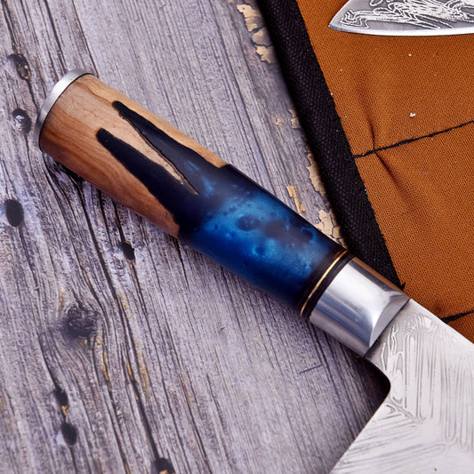 A close-up of a knife handle with a natural wood top, a blue marbled resin section, and a polished silver bolster, resting on a weathered gray wooden surface next to a brown fabric sheath.