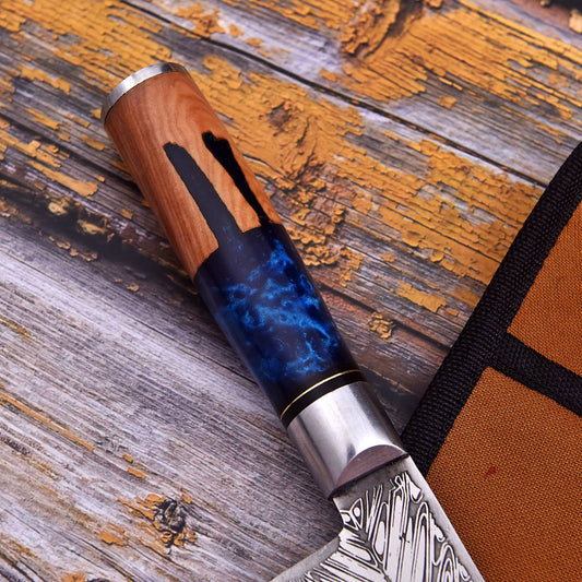 A close-up of a knife with a patterned steel blade, a silver bolster, and a wooden handle featuring a blue marbled inlay, resting on a weathered wooden surface next to an orange fabric sheath.