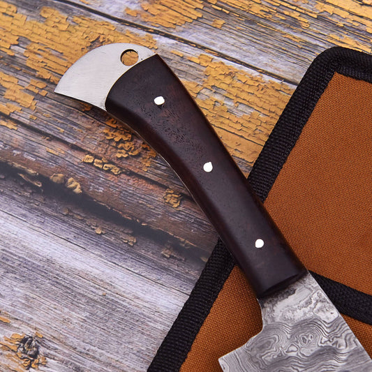 A close-up of a handcrafted knife with a dark brown wooden handle and a patterned steel blade, resting on a rustic weathered wood surface next to its orange and black sheath.