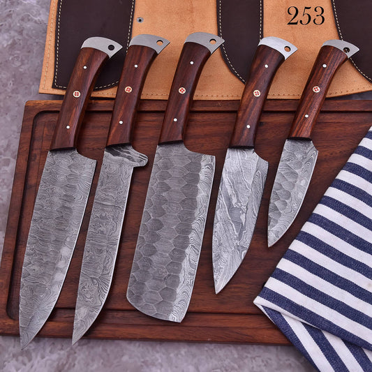 Five Damascus steel kitchen knives with dark wood handles are arranged on a wooden cutting board, with a tan knife sheath and a navy-and-white striped cloth visible in the background.