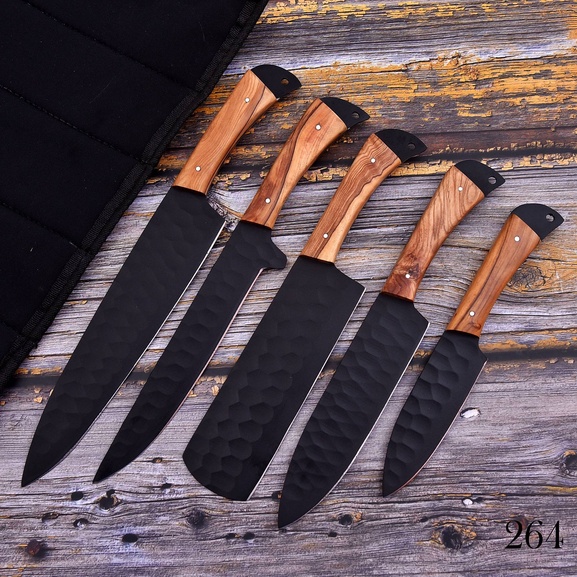 Five kitchen knives with black hammered blades and light brown wood handles are arranged diagonally on a rustic weathered wood surface next to a black carrying case.