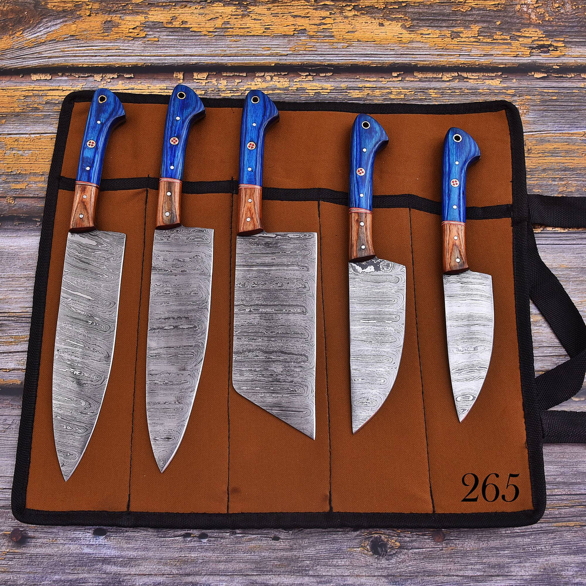 Five kitchen knives with patterned steel blades and blue handles are displayed in a brown knife roll on a rustic wooden surface. The knife roll has black trim and the number 265 printed in black in the bottom right corner.