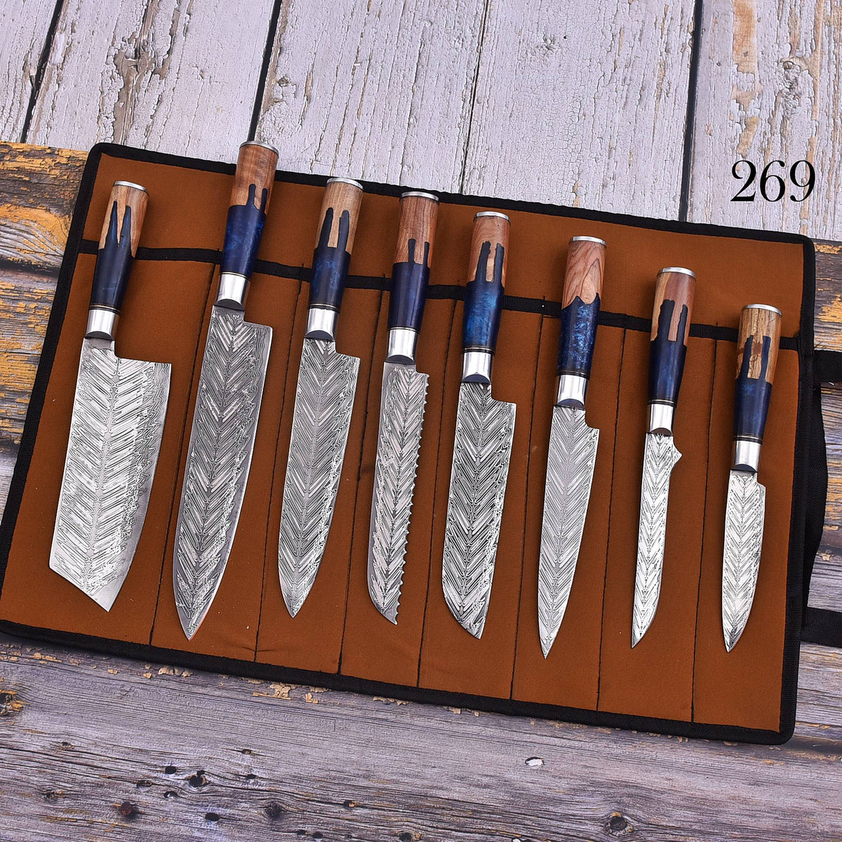 A set of eight kitchen knives with patterned Damascus steel blades and dark blue handles, displayed in a brown roll-up leather case on a rustic wooden surface. The number '269' is printed in black in the top right corner.