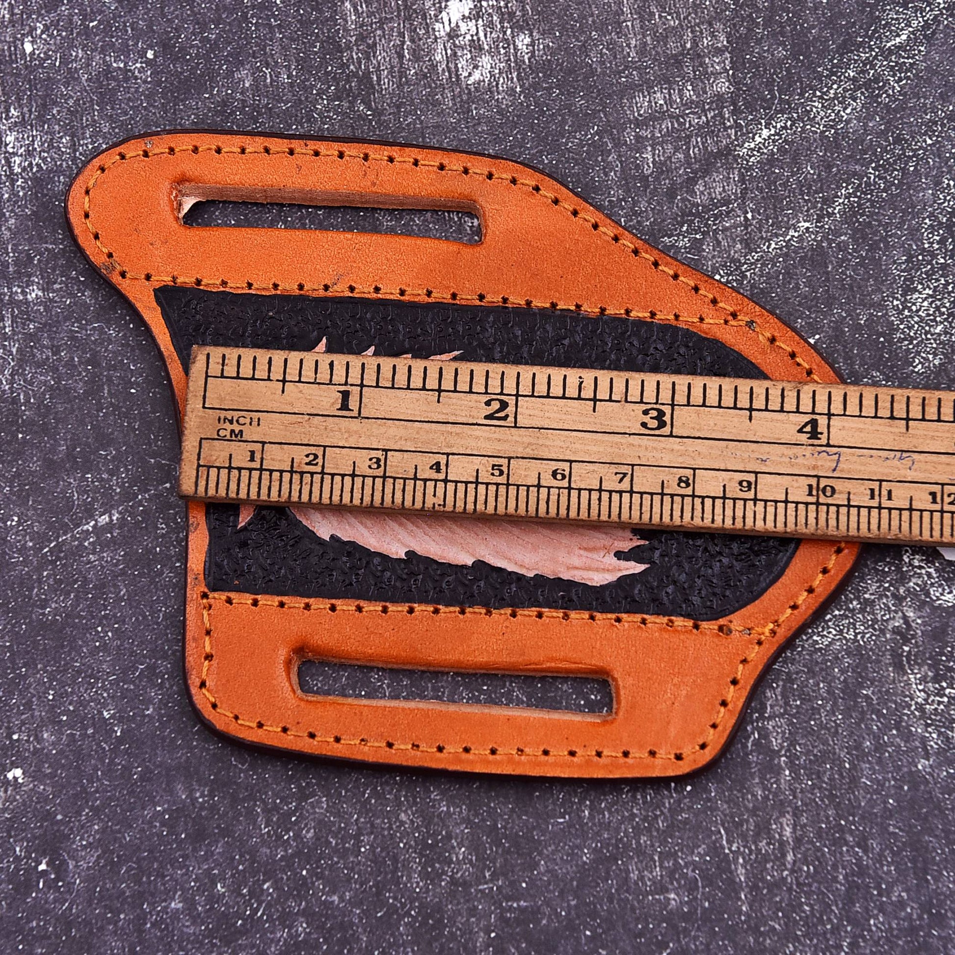 A tan leather belt holster or sheath with two cutout slots, shown on a dark textured surface with a wooden ruler placed across the center to indicate size.