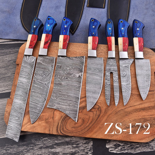 A 7-piece kitchen knife set with patterned Damascus steel blades and patriotic red, white, and blue handles, displayed on a wooden cutting board with a blue leather sheath in the background.