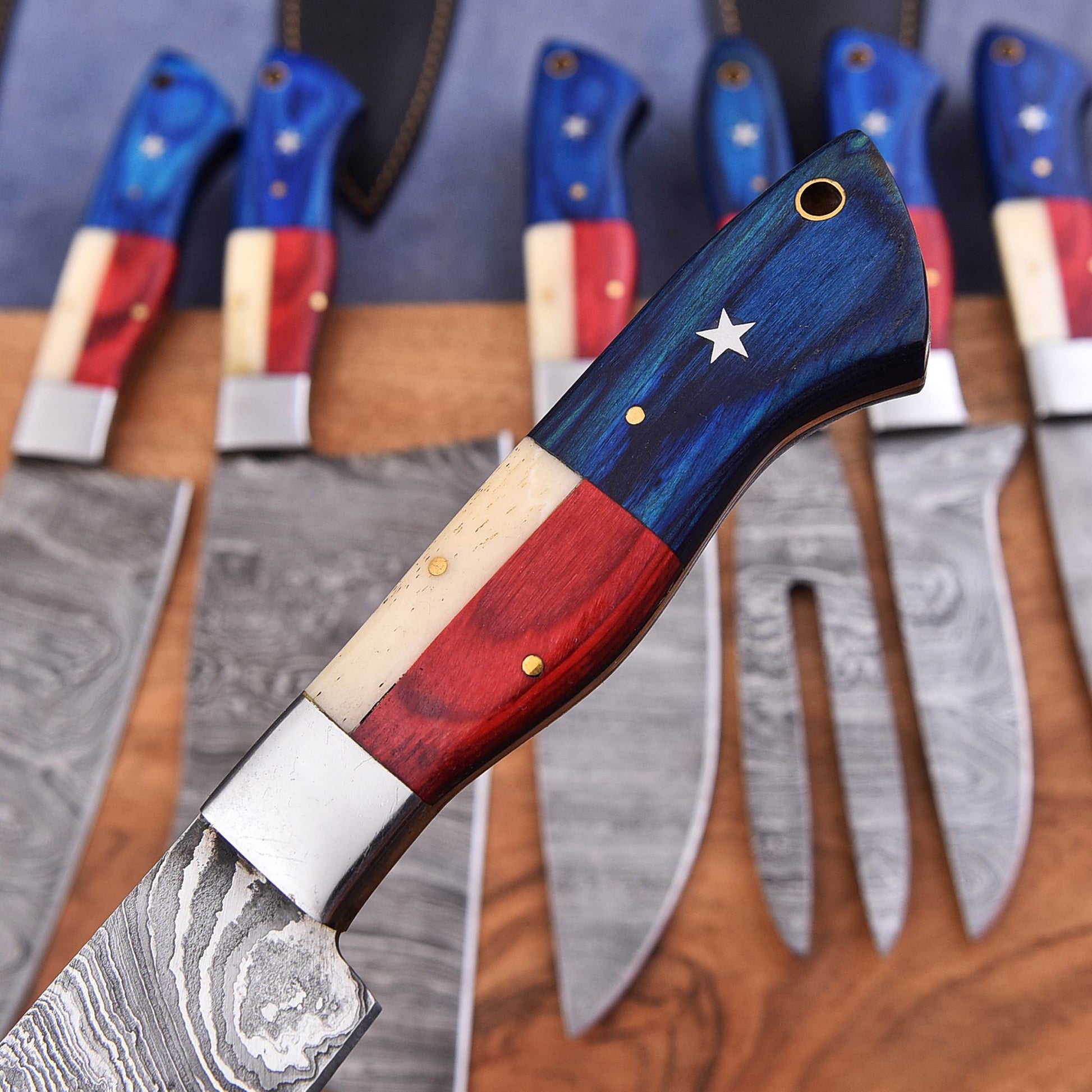 A close-up of a decorative chef's knife with a handle painted like the American flag, with other knives in the background on a wooden surface.