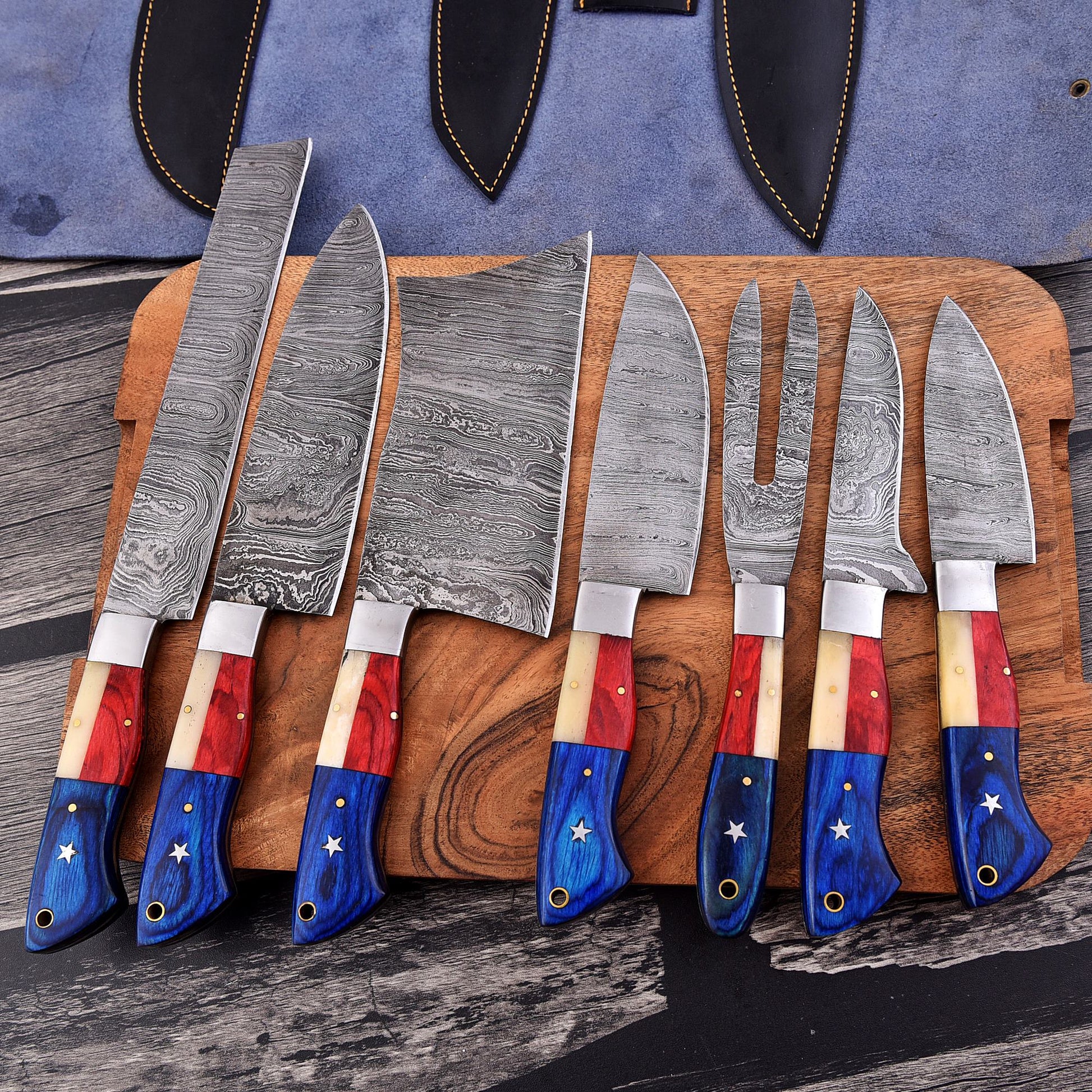 A set of seven Damascus steel kitchen knives with patriotic red, white, and blue handles, arranged on a wooden cutting board with a blue leather sheath in the background.