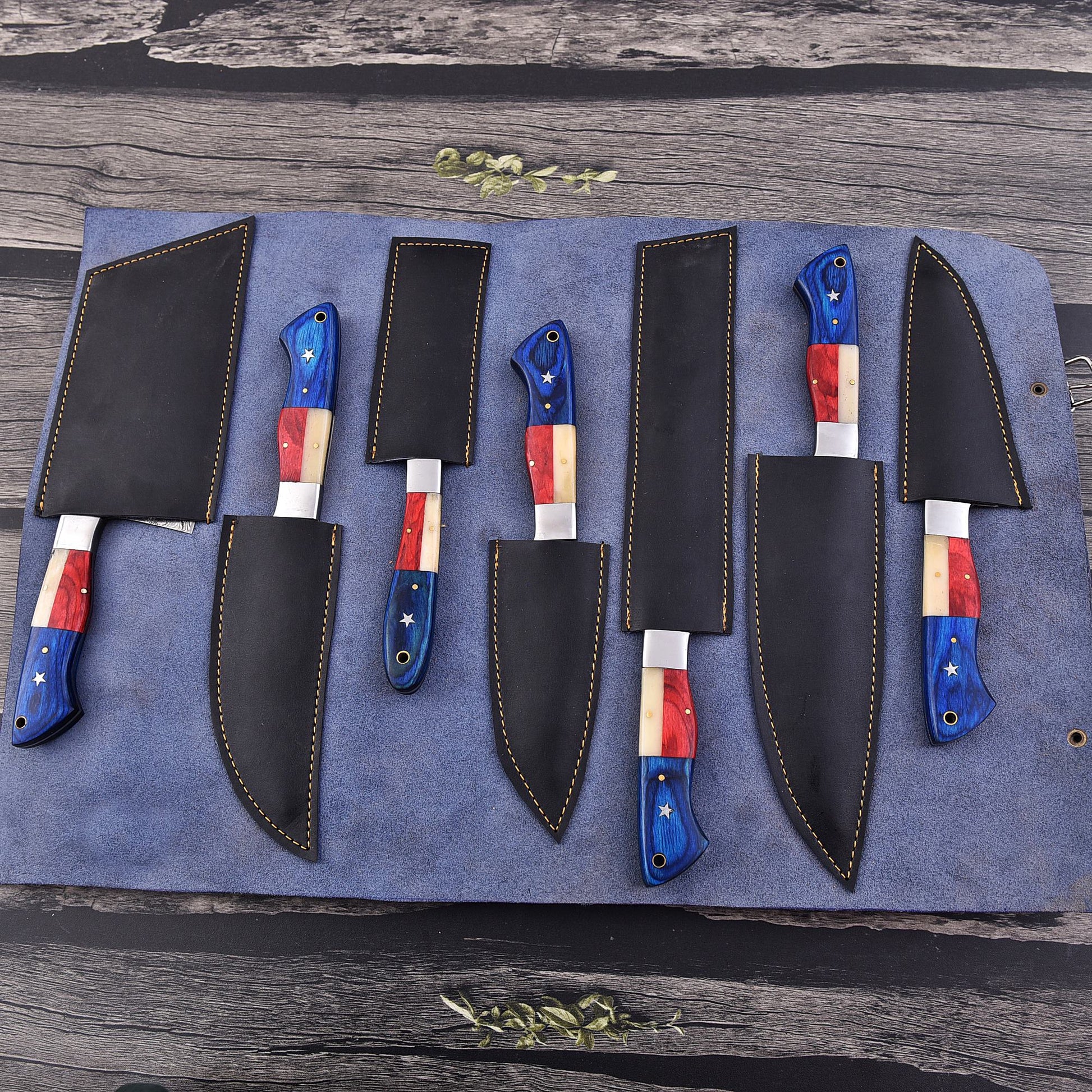 A set of six kitchen knives with black leather sheaths arranged on a blue fabric roll over a rustic wooden surface. The knife handles feature a red, white, and blue flag-inspired pattern with small white star details.
