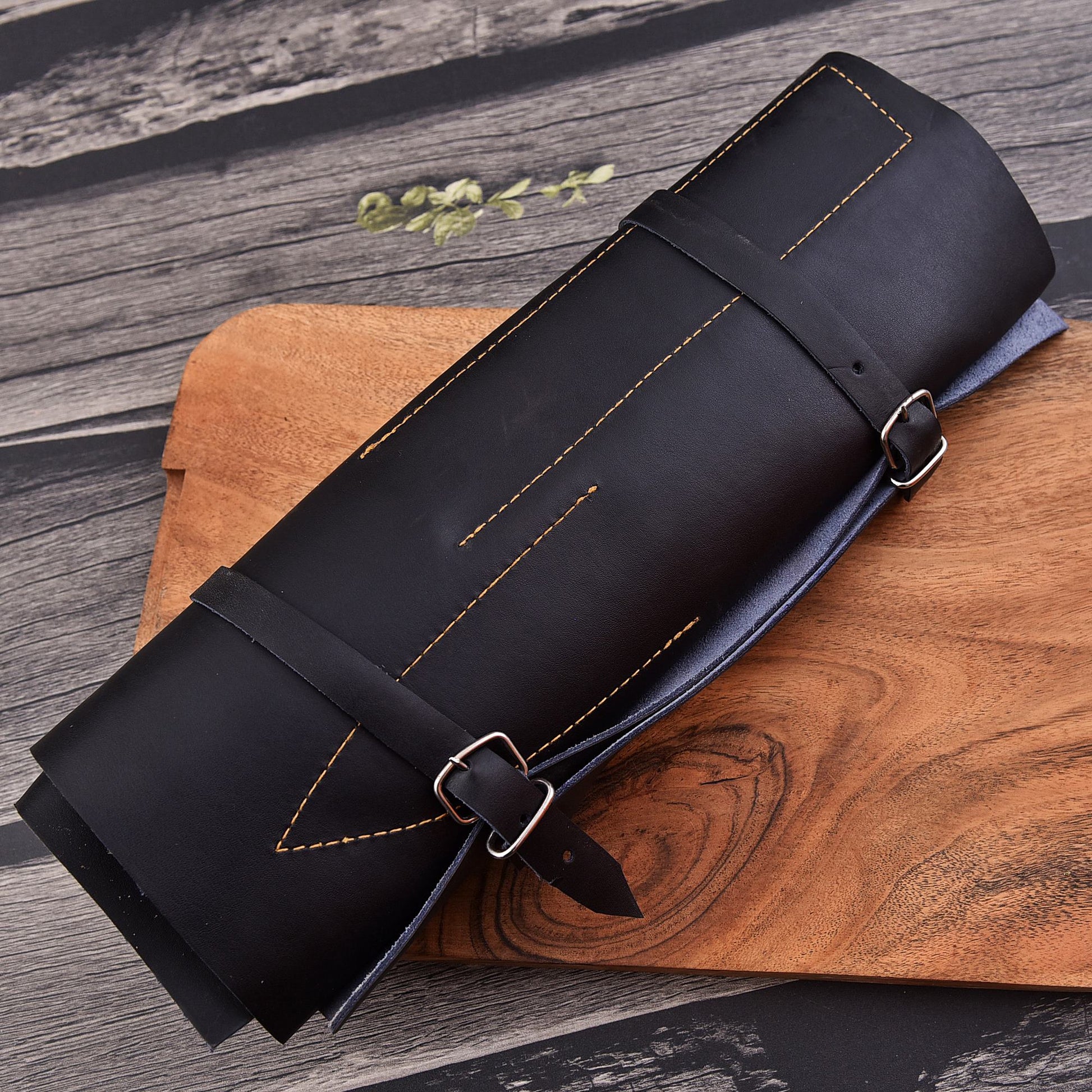A black leather rolled mat or towel with gold contrast stitching and buckle straps, resting on a wooden cutting board over a dark wood surface.