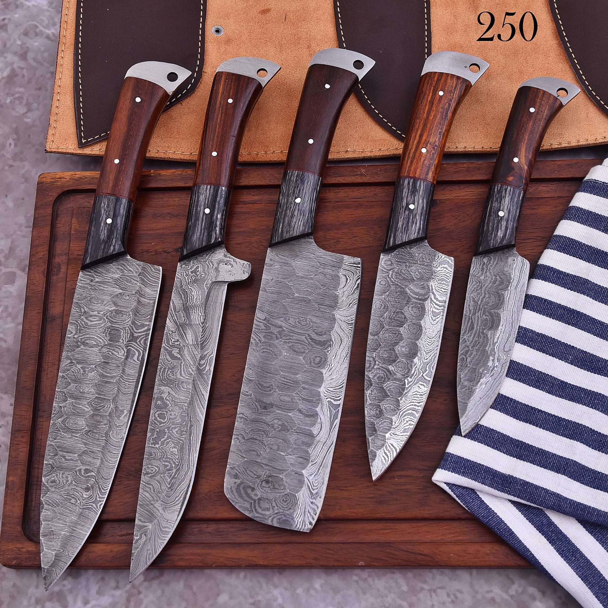 Five kitchen knives with patterned Damascus steel blades and dark wood handles are arranged on a wooden cutting board, with a tan leather sheath and a striped cloth visible in the background.
