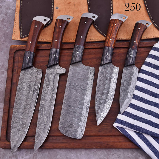 Five kitchen knives with patterned Damascus steel blades and dark wood handles are arranged on a wooden cutting board, with a tan leather sheath and a striped cloth visible in the background.