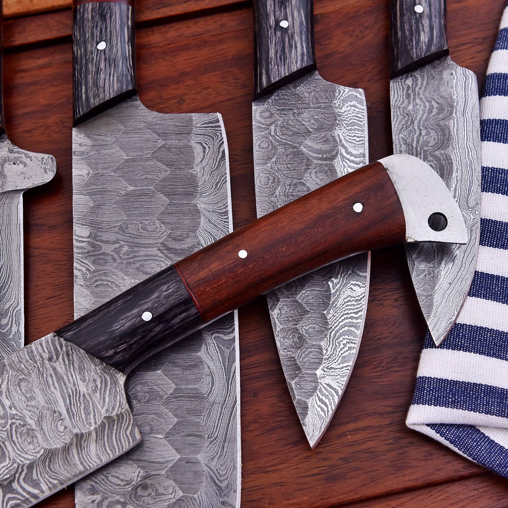 A set of kitchen knives with patterned Damascus steel blades and dark wood handles, arranged on a dark wooden surface next to a blue and white striped cloth.