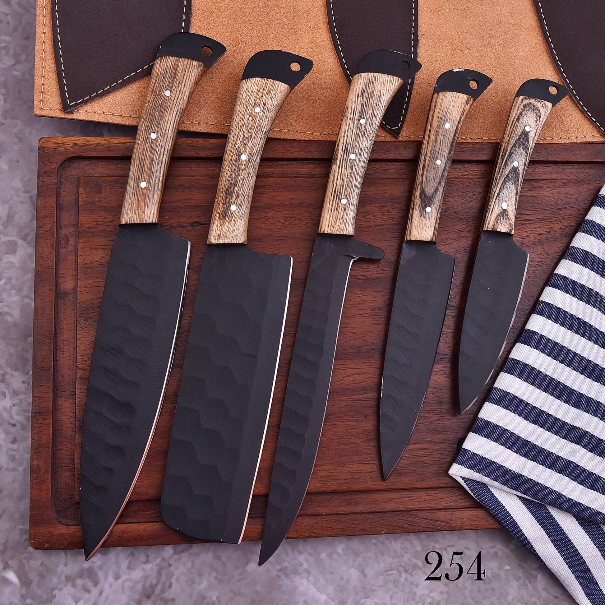 Five-piece handmade chef knife set with black hammered blades, wooden handles and leather blade covers on a wooden cutting board