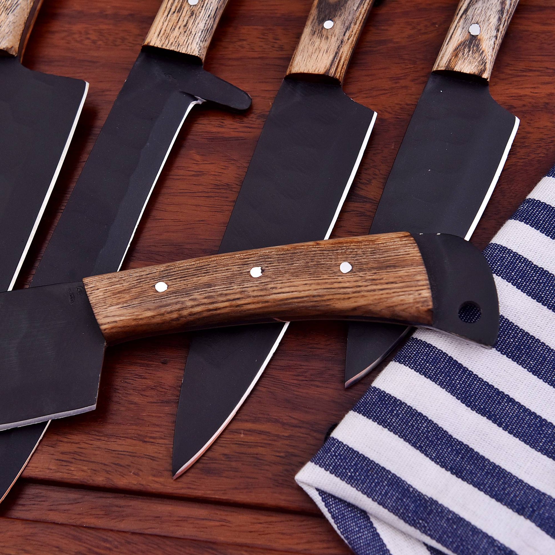 A set of large kitchen knives with dark steel blades and brown wood-grain handles, arranged diagonally on a dark wooden surface next to a blue and white striped cloth.