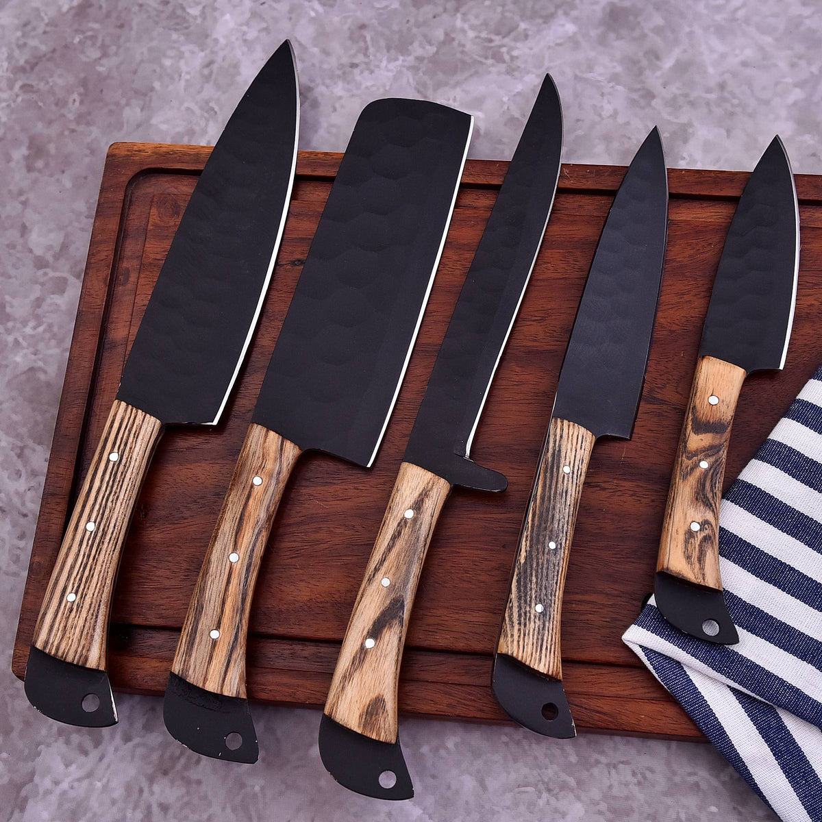 Five kitchen knives with black hammered blades and brown wood-grain handles arranged on a dark wooden cutting board, with a striped cloth partially visible at the corner.