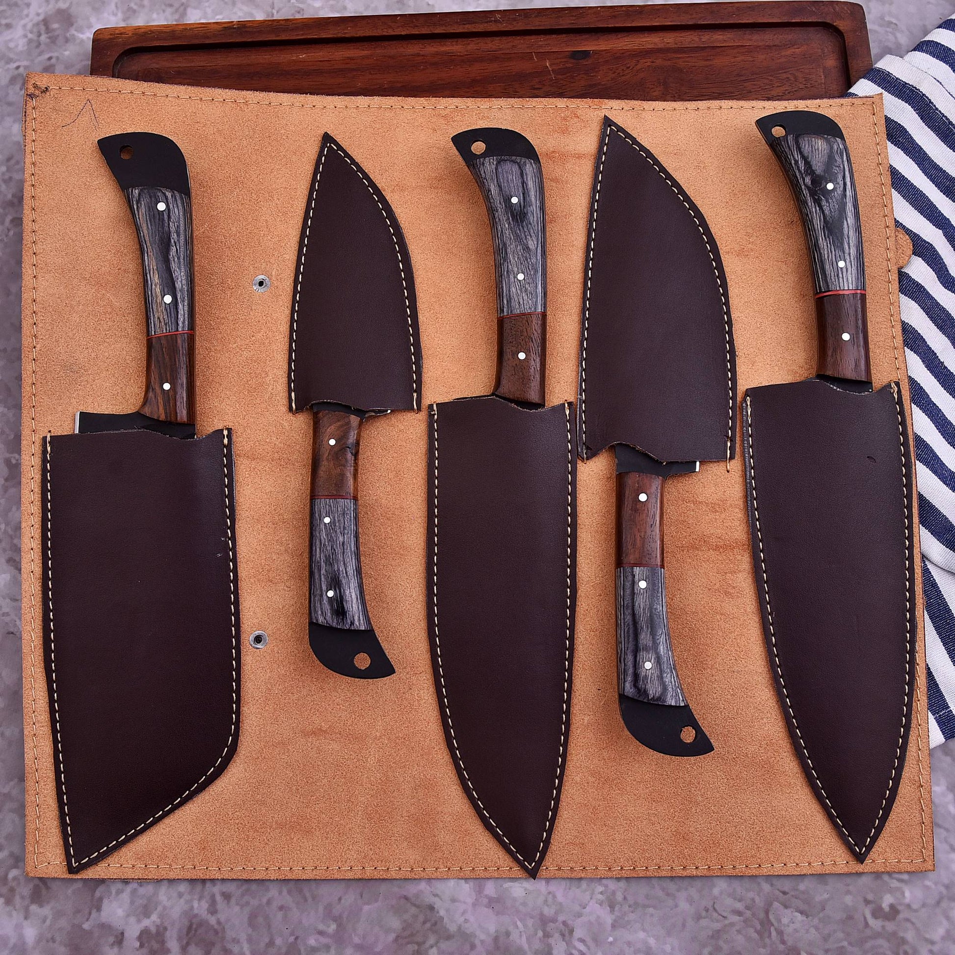 A set of five kitchen knives with dark wood and gray marbled handles, displayed in a tan leather roll-up sheath on a wooden surface.