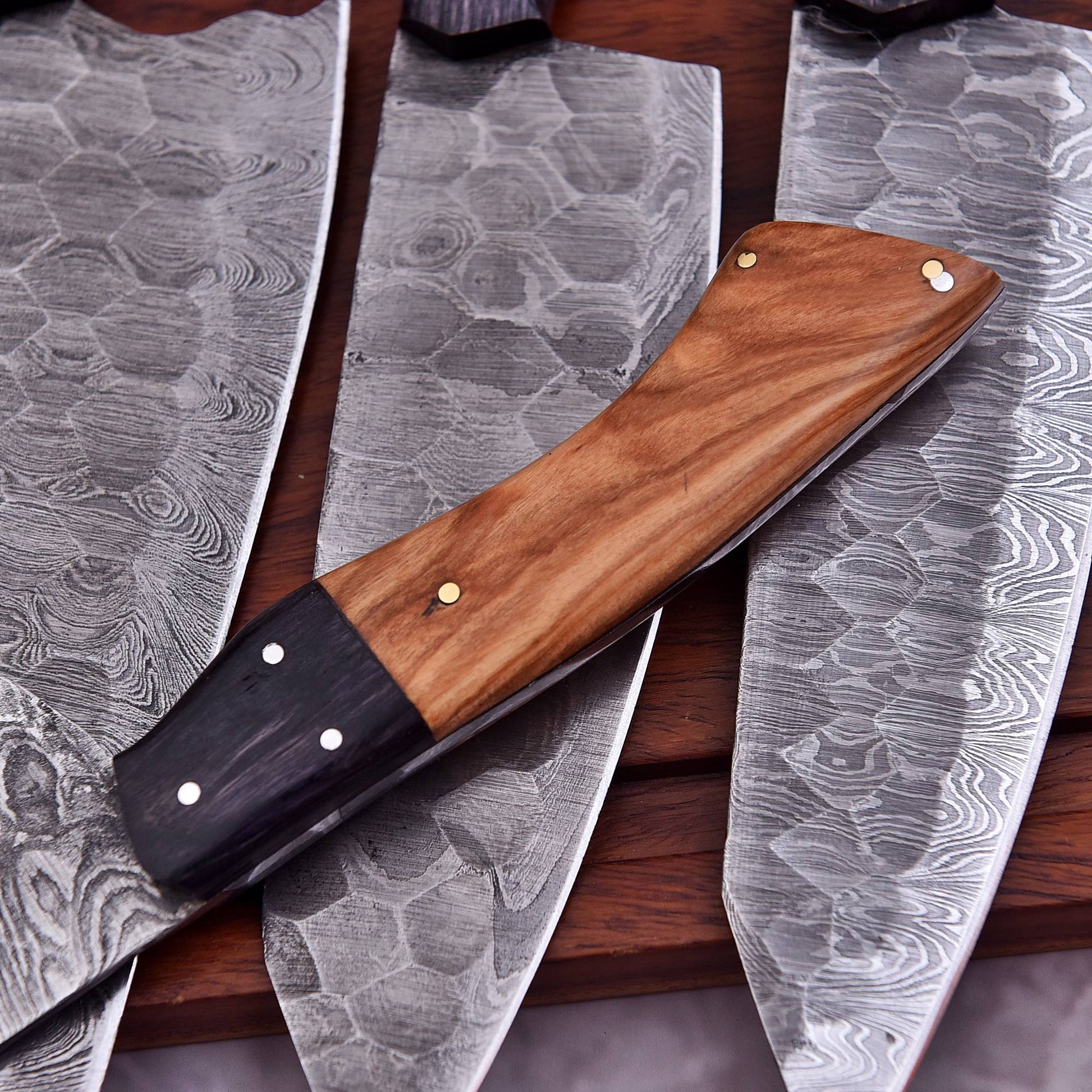 A close-up of a handmade chef's knife with a patterned Damascus steel blade and a wooden handle, laid diagonally over other similar knives on a dark wooden surface.