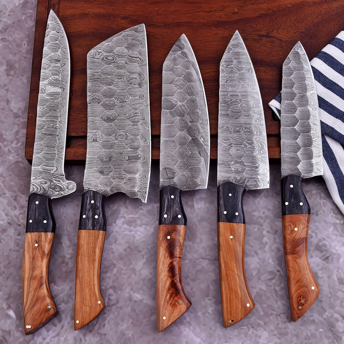 Five kitchen knives with patterned steel blades and wooden handles arranged on a dark wooden board over a gray textured surface, with a striped cloth partially visible on the right.