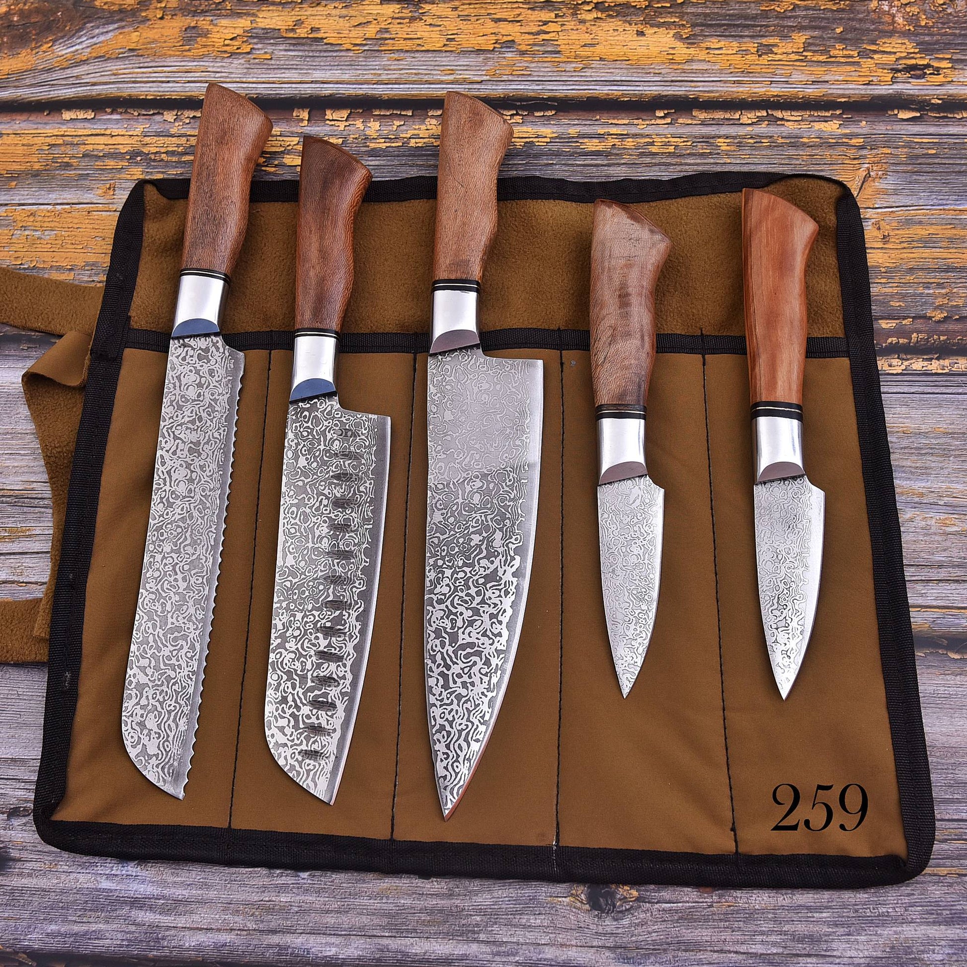 A set of five kitchen knives with patterned steel blades and brown wooden handles, displayed in a brown fabric knife roll on a rustic wooden surface.