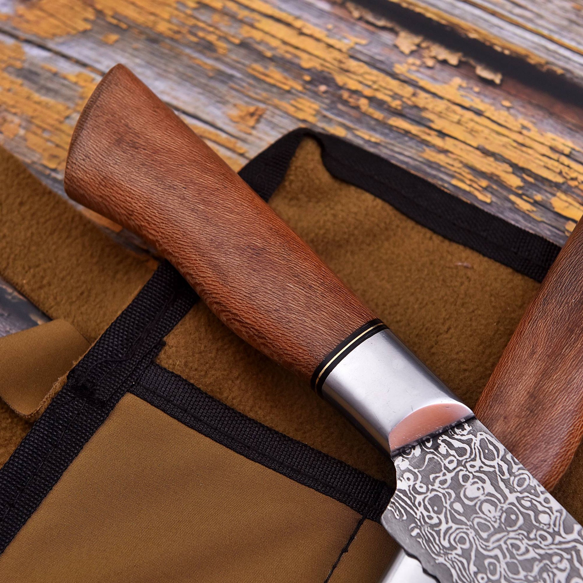 A close-up of a Damascus steel kitchen knife with a brown wooden handle resting on a brown leather sheath with black trim, set against a rustic wooden background.