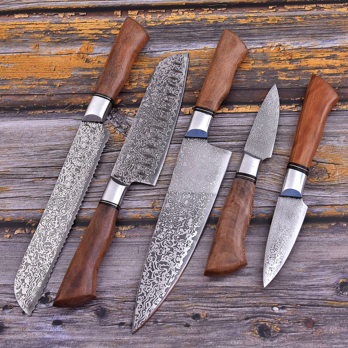 Five Damascus steel kitchen knives with brown wooden handles arranged diagonally on a rustic, weathered wooden surface.