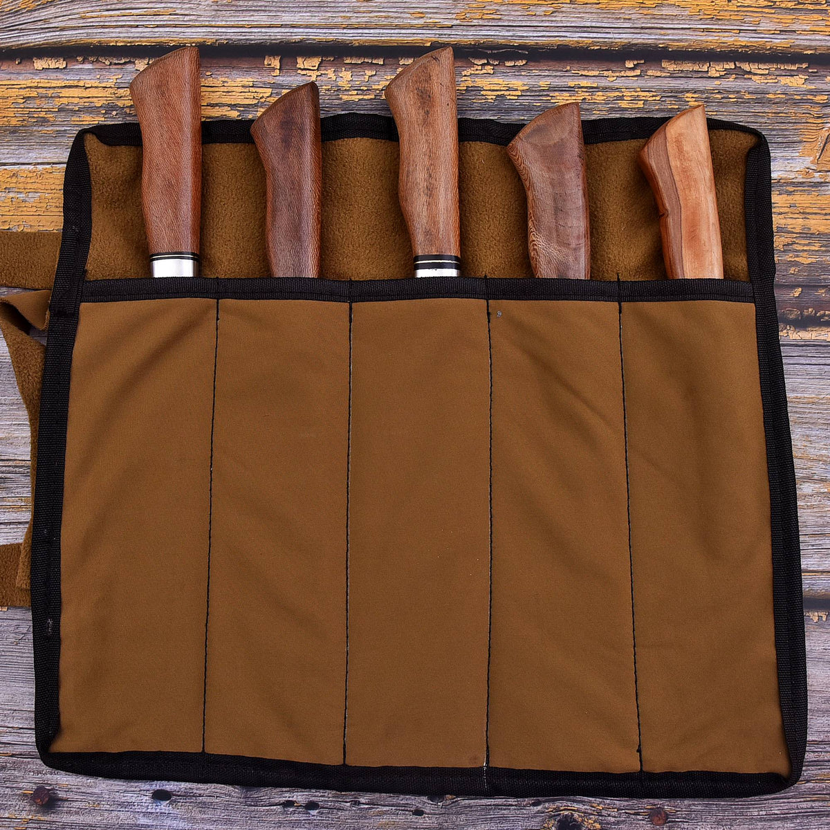 A brown fabric knife roll with black trim holding five knives with wooden handles, laid flat on a rustic wooden surface.