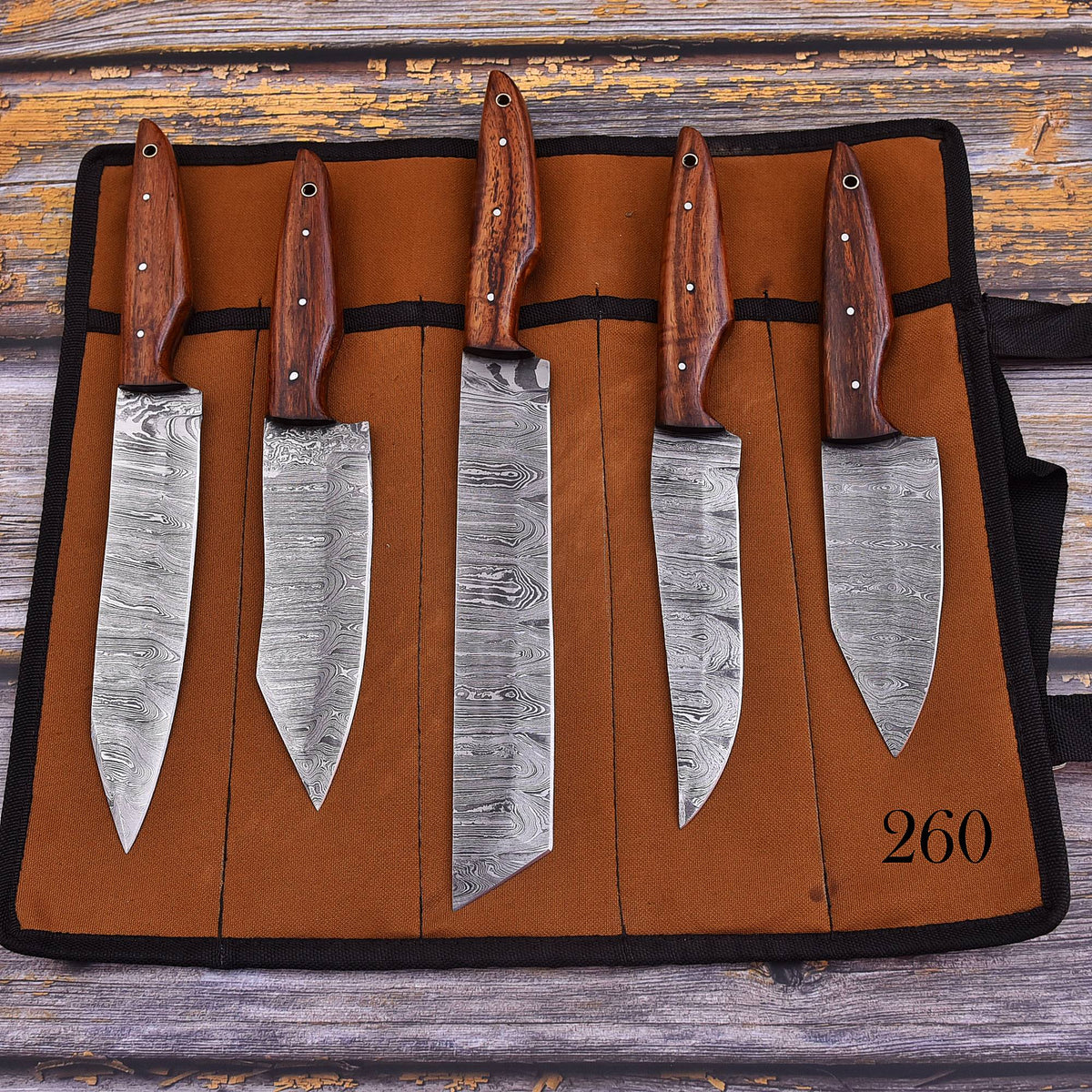 A set of five kitchen knives with patterned steel blades and brown wooden handles, displayed in a brown roll-up knife case on a rustic wooden surface.