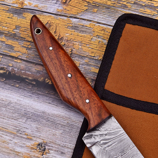 A close-up of a knife with a polished reddish-brown wooden handle and a patterned steel blade, resting diagonally on a rustic weathered wood surface next to an orange and black fabric sheath.