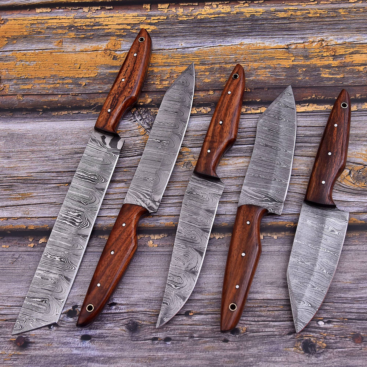 Five fixed-blade knives with patterned steel blades and dark brown wooden handles arranged diagonally on a rustic weathered wood background.