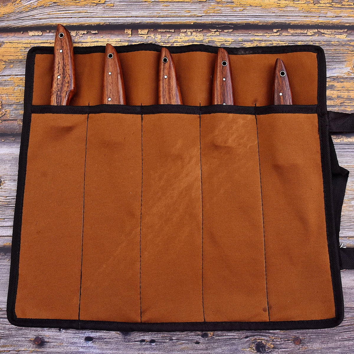 A brown canvas knife roll with black trim holding five knives with brown wooden handles, laid flat on a rustic wooden surface.