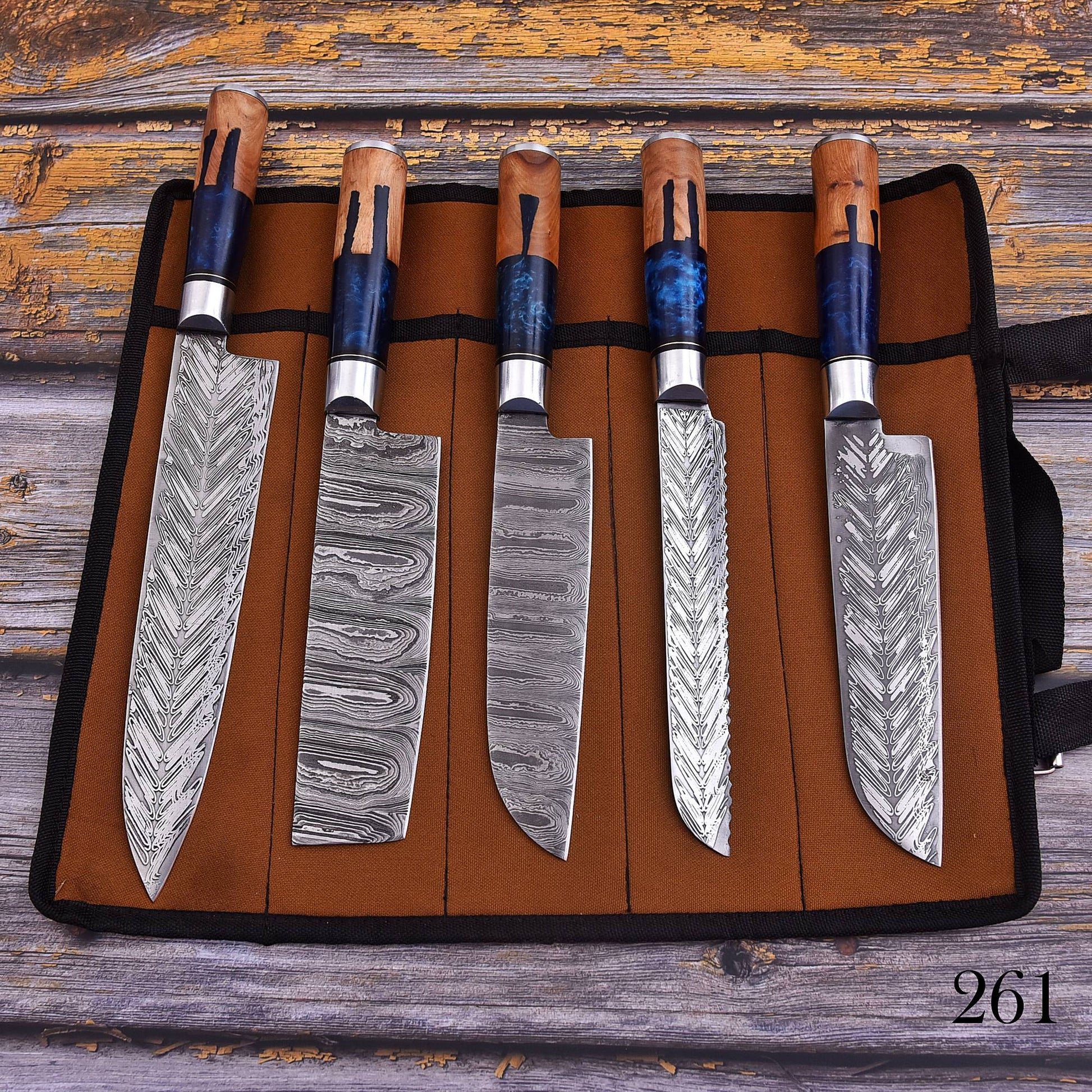 Five kitchen knives with patterned Damascus steel blades and wooden handles are displayed in a brown knife roll on a rustic wooden surface.