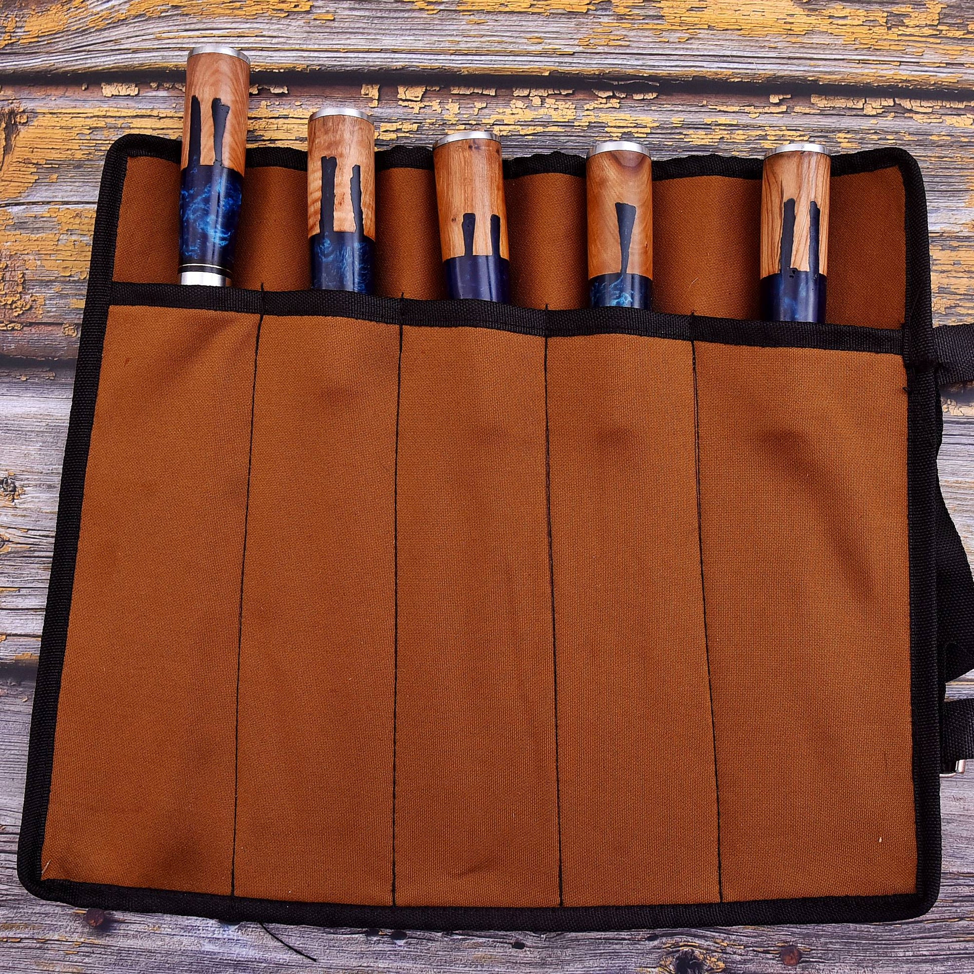 A brown fabric roll-up case with black trim holding five wooden-handled tools, photographed from above on a rustic weathered wood surface.