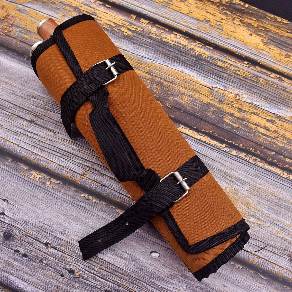 A rolled tan canvas or leather-like protective sleeve with black straps and silver buckle hardware, placed diagonally on a weathered wooden surface.