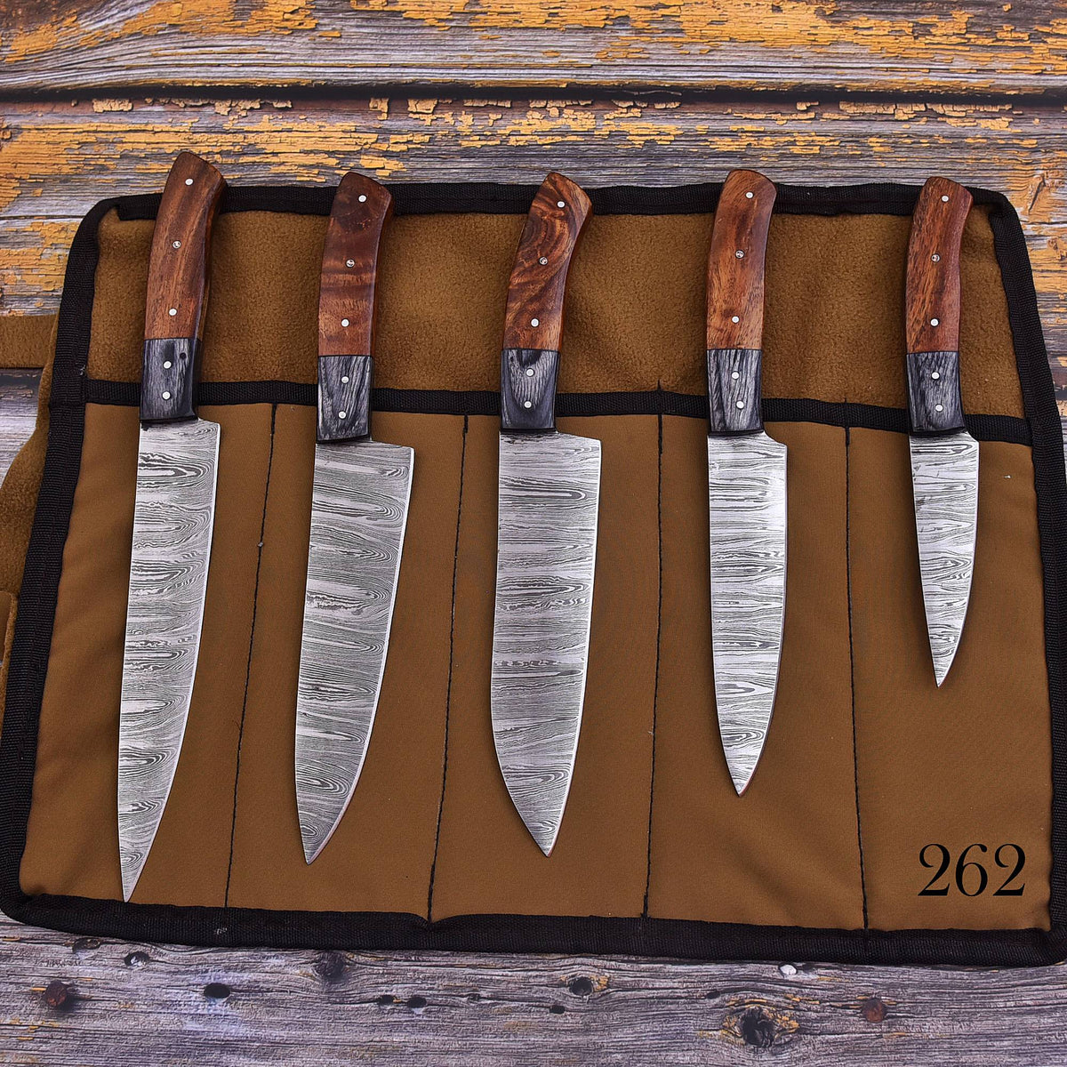 Five kitchen knives with patterned steel blades and wooden handles are displayed in a brown fabric knife roll on a rustic wooden surface. The roll has black trim and the number 262 printed in black in the bottom right corner.