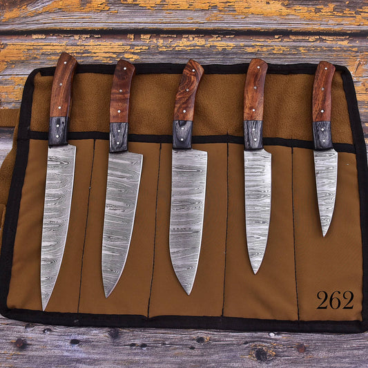 Five kitchen knives with patterned steel blades and wooden handles are displayed in a brown fabric knife roll on a rustic wooden surface. The roll has black trim and the number 262 printed in black in the bottom right corner.