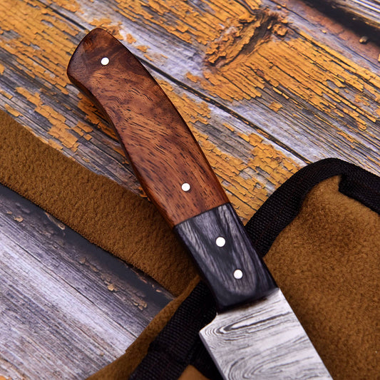 A close-up of a knife with a dark wood handle and Damascus steel blade, resting on a brown leather sheath over a rustic weathered wood background.