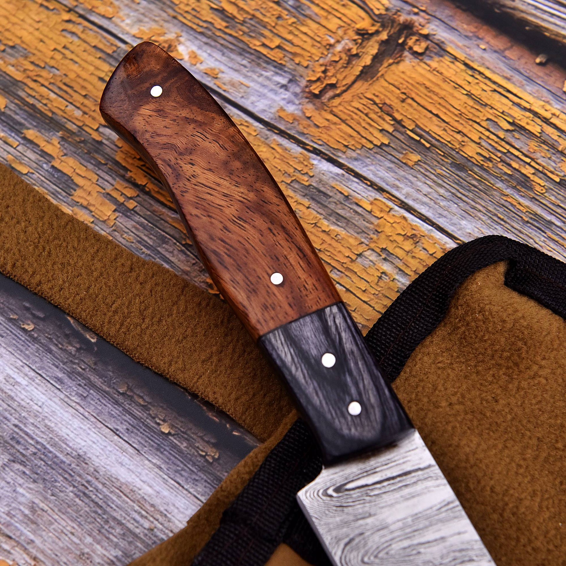 A close-up of a knife with a dark wood handle and Damascus steel blade, resting on a brown leather sheath over a rustic weathered wood background.