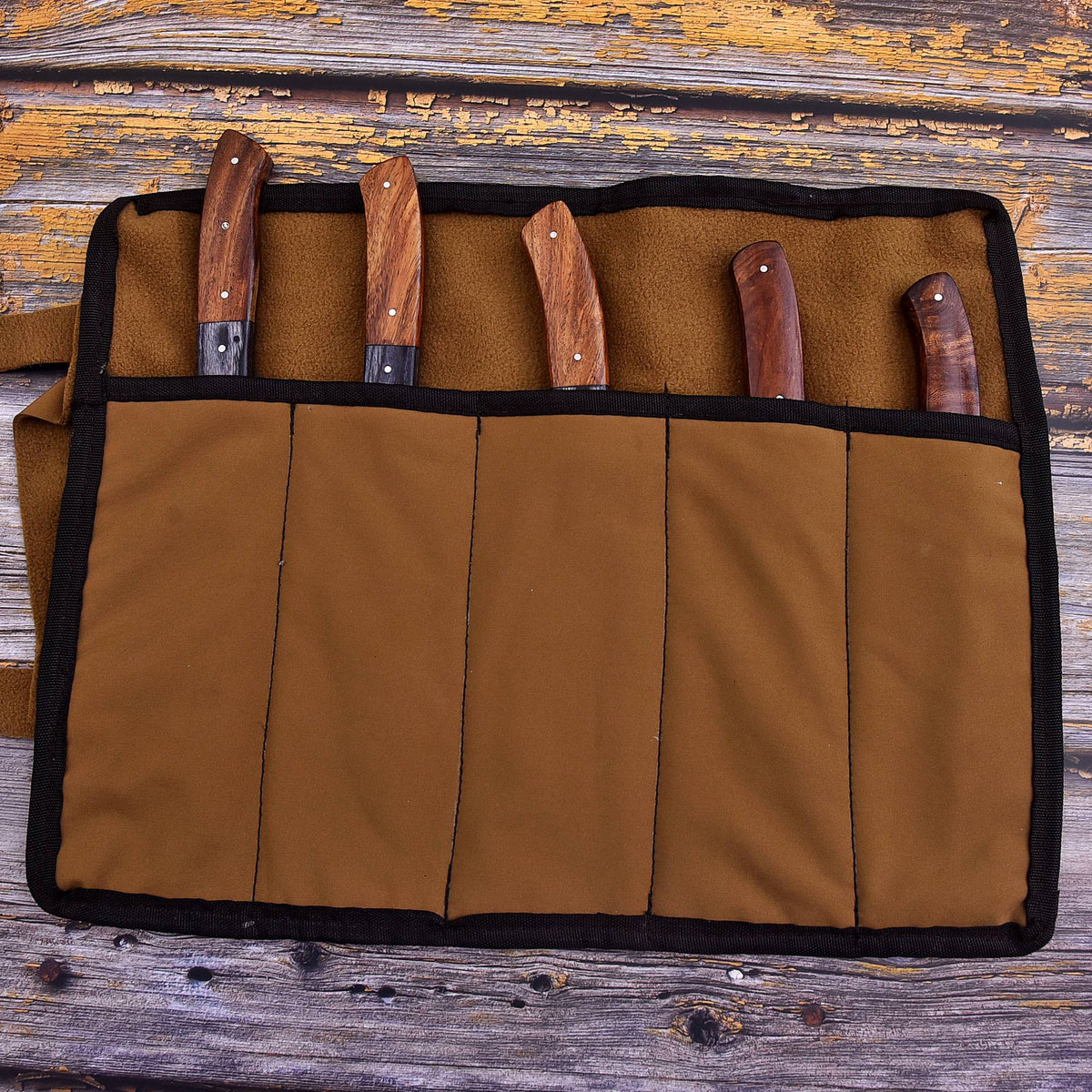 A brown canvas knife roll with black trim holding five knives with wooden handles, laid flat on a rustic weathered wood background.