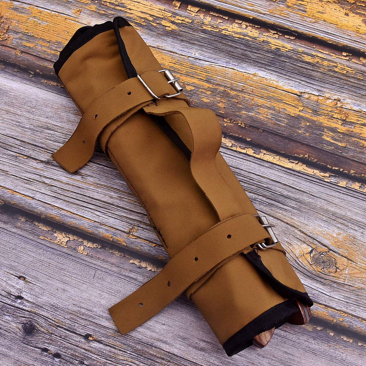 A rolled tan leather or canvas arm guard with multiple adjustable straps and silver-tone metal buckles, photographed on a rustic weathered wood surface.
