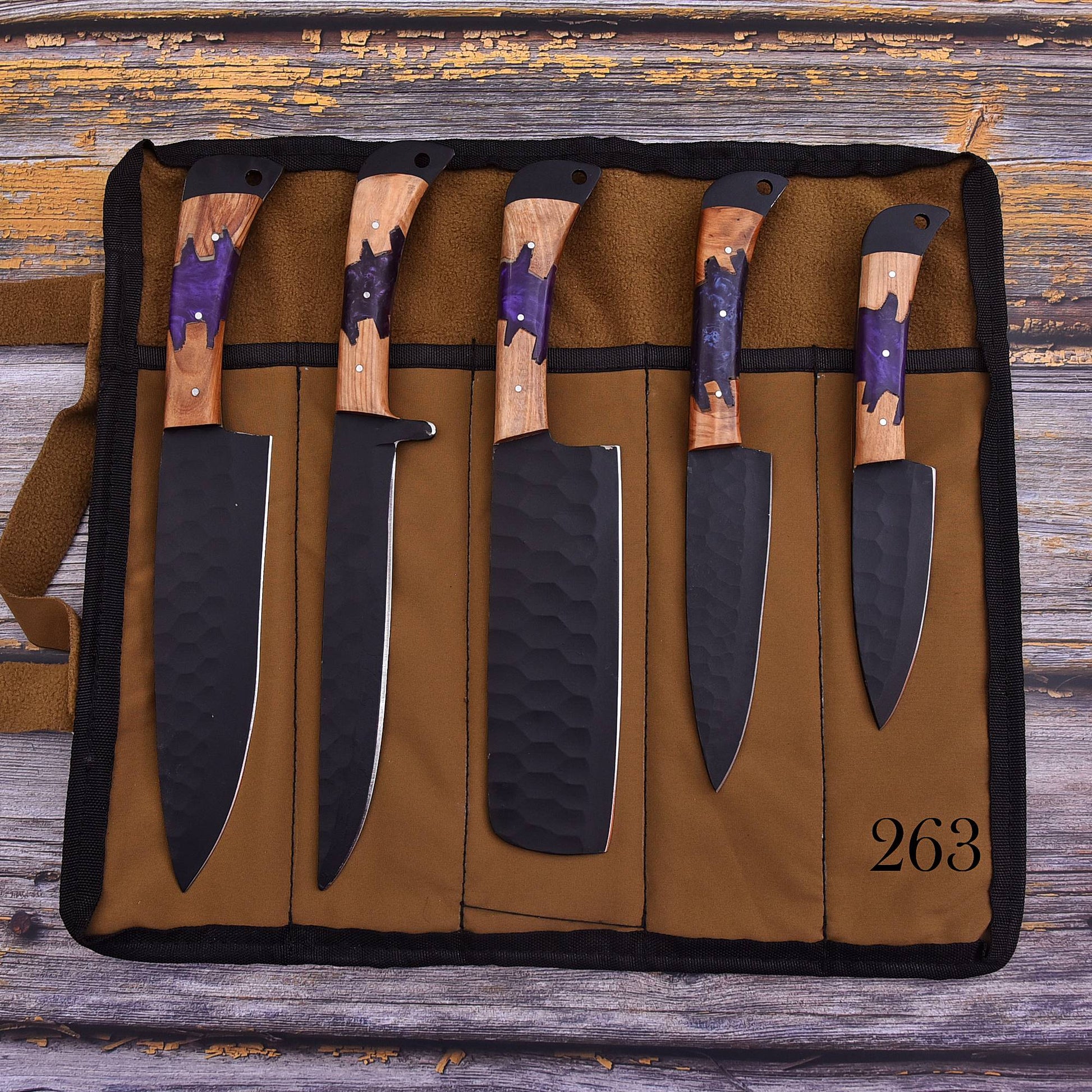 A set of five kitchen knives displayed in a brown canvas roll-up knife case on a rustic wooden surface. The knives have black hammered blades and wood handles with purple inlay.