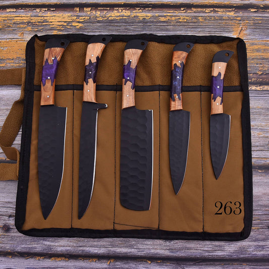 A set of five kitchen knives displayed in a brown canvas roll-up knife case on a rustic wooden surface. The knives have black hammered blades and wood handles with purple inlay.