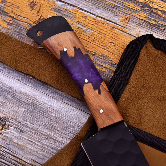 A close-up of a custom chef's knife with a wooden handle featuring a purple painted cityscape design and silver rivets, resting on a brown fleece sheath over a rustic wooden background.