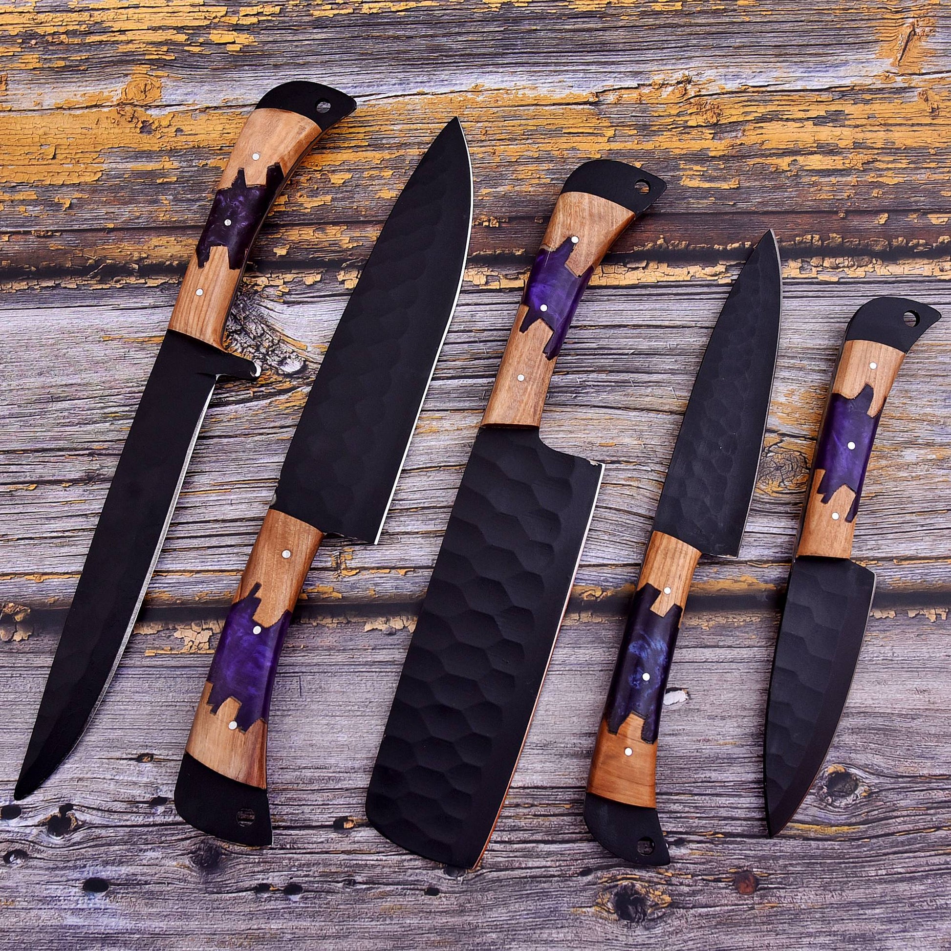 Four kitchen knives with black hammered blades and purple-and-wood handles, arranged diagonally on a rustic weathered wood surface.
