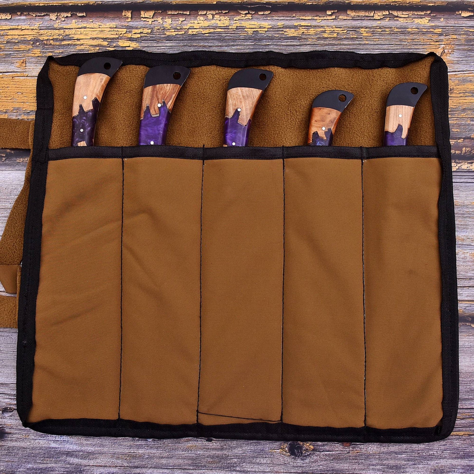 A brown canvas knife roll with black trim holding five knives with wooden and purple handles, laid flat on a rustic wooden surface.