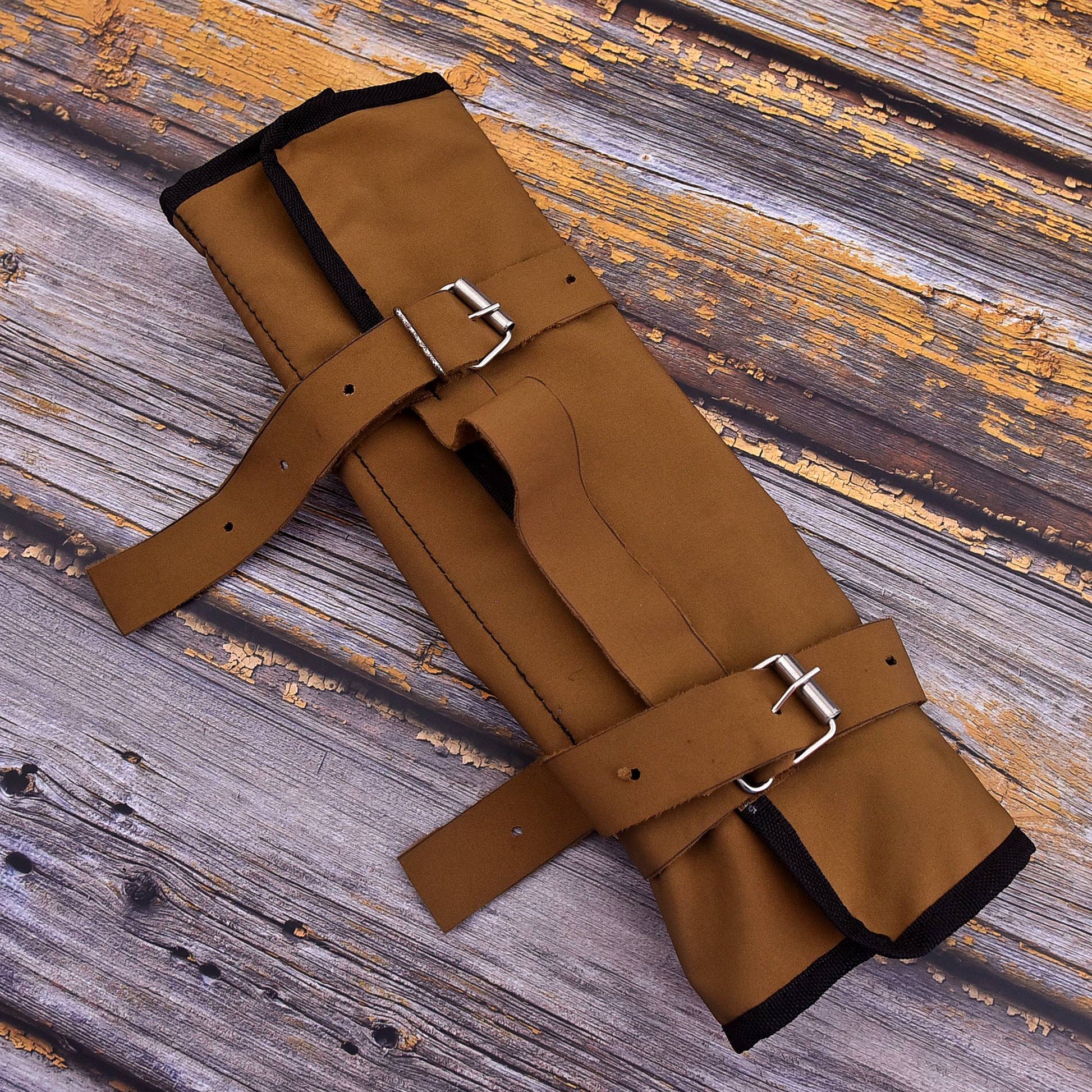 A brown leather bracer or guard with black trim and silver metal buckles, laid flat on a rustic wooden plank background.