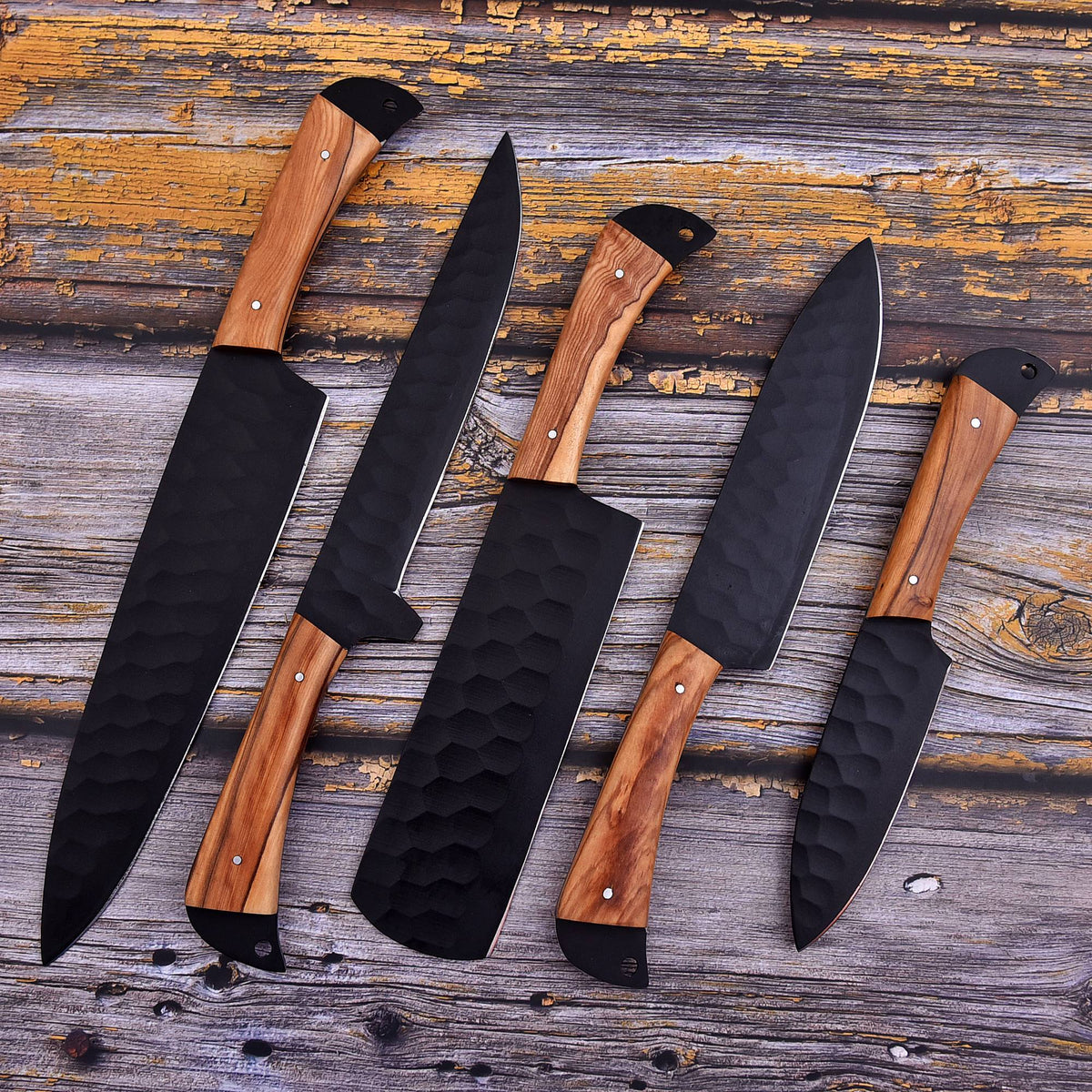 Five kitchen knives with black hammered blades and light brown wooden handles arranged diagonally on a rustic weathered wood surface.