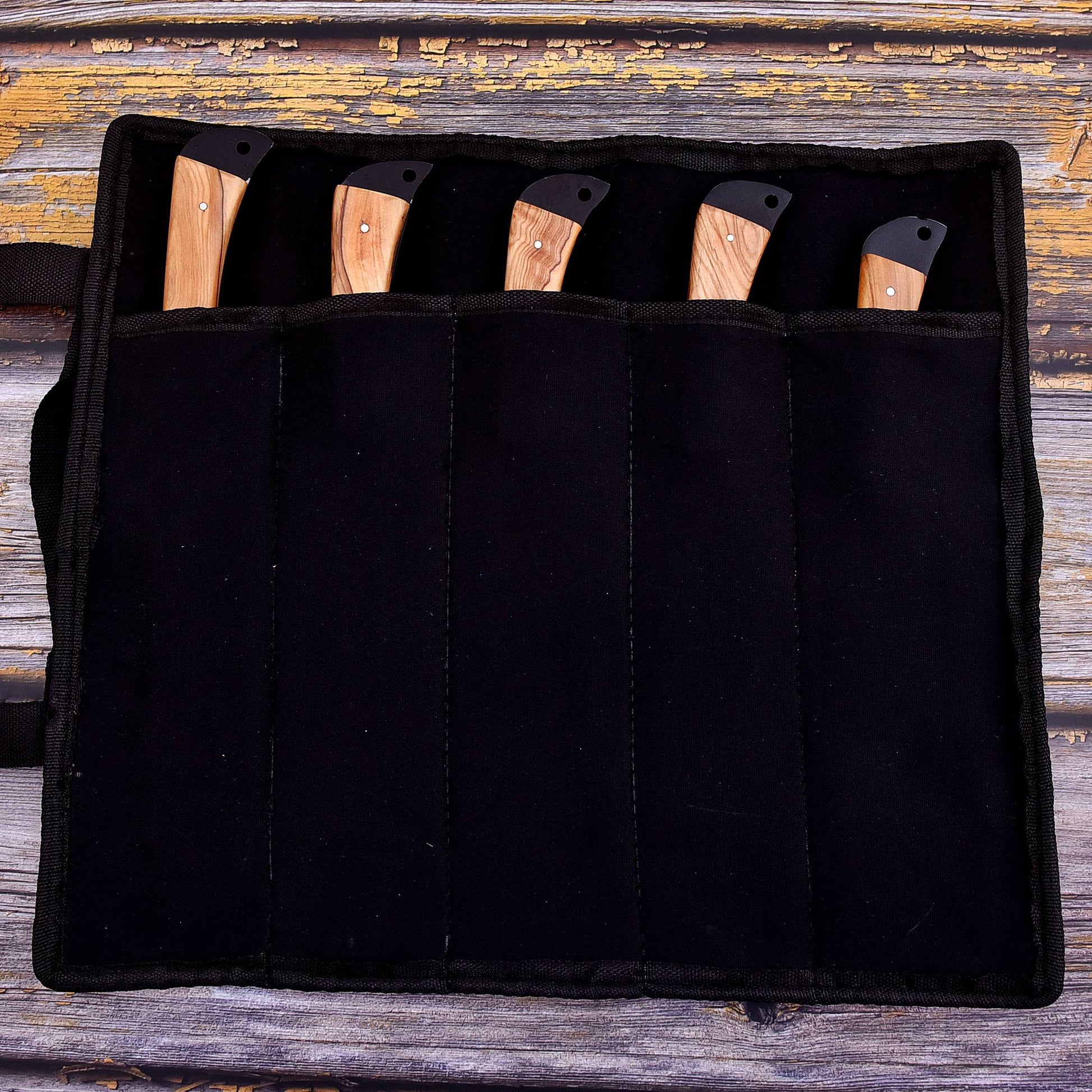 A black fabric knife roll holding five knives with light wood handles and black blades, laid flat on a weathered wooden surface.