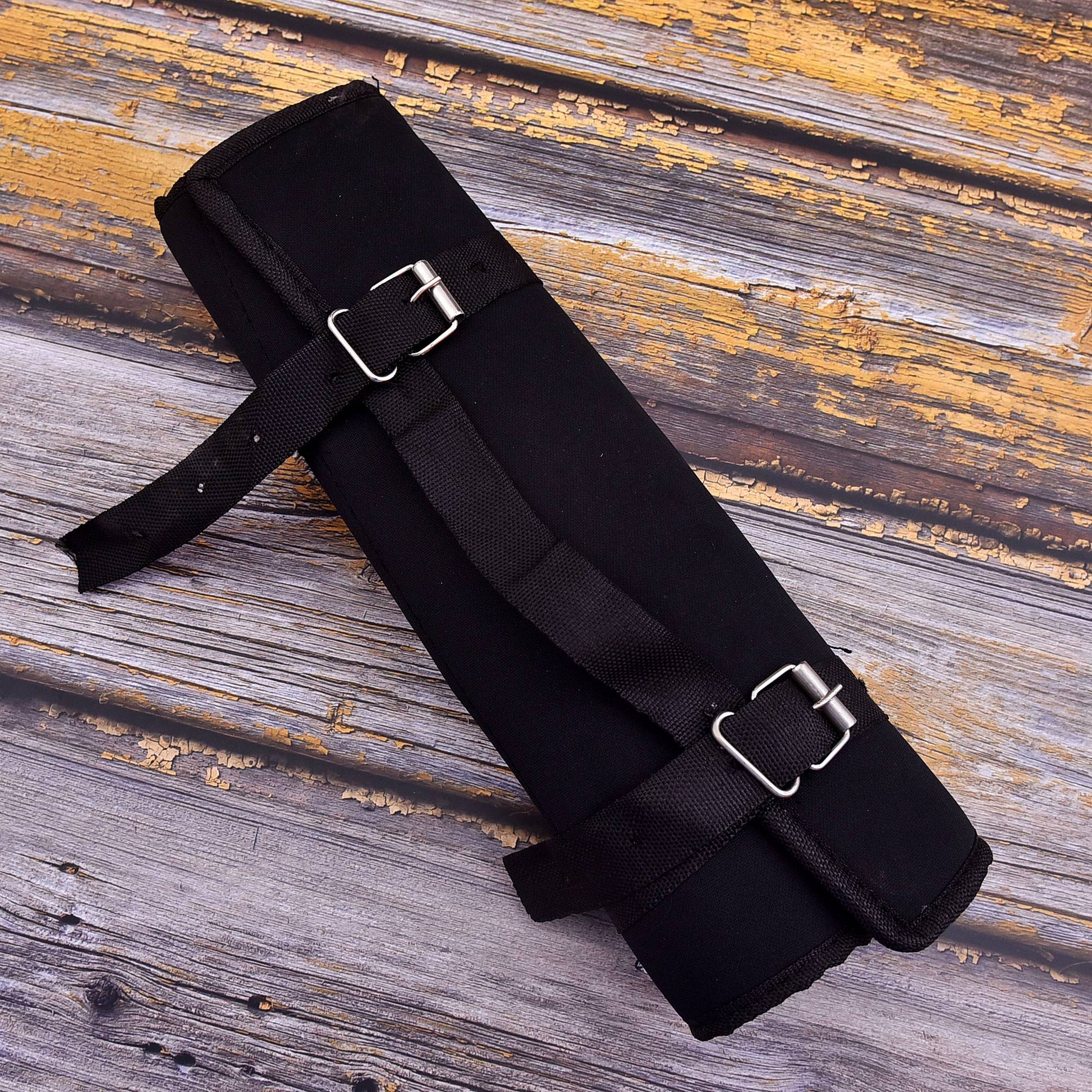 A black padded cylindrical roll or sleeve with two adjustable black straps and silver metal buckles, photographed on a weathered wood surface.