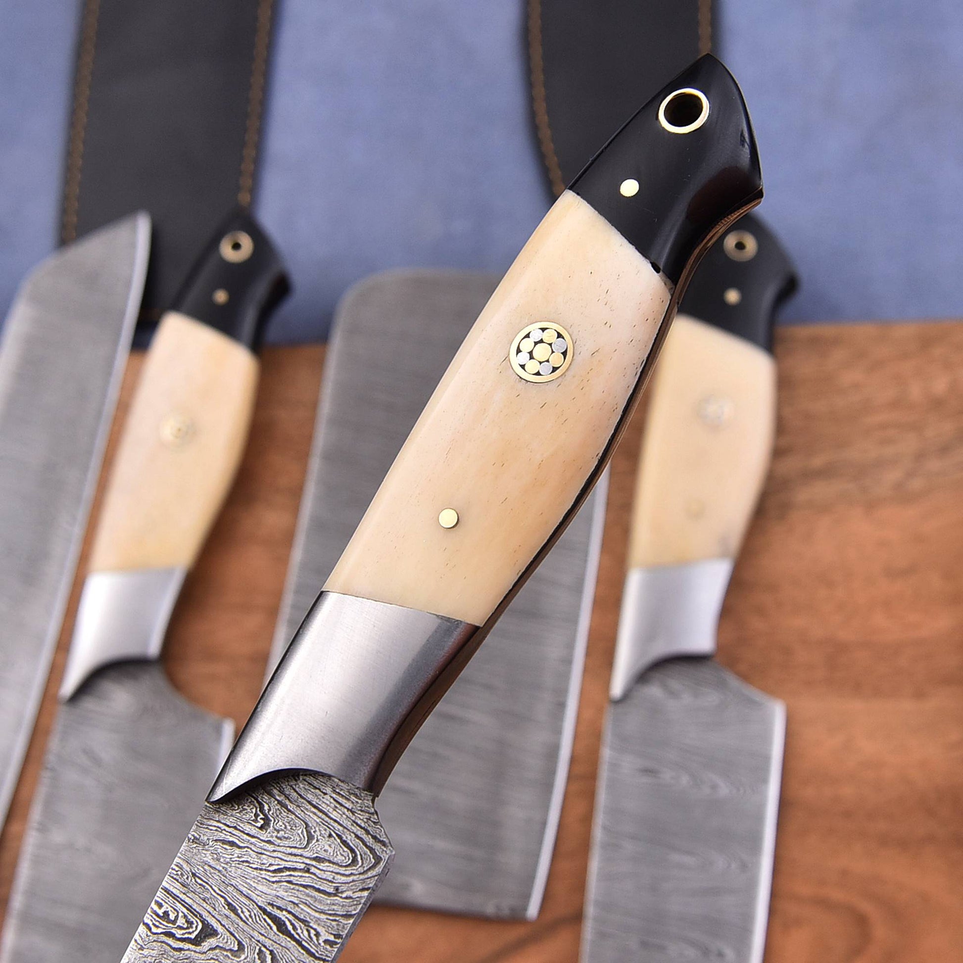 A close-up of a Damascus steel chef's knife with a cream-colored bone handle and brass pins, displayed in front of a matching set of kitchen knives on a wooden surface.