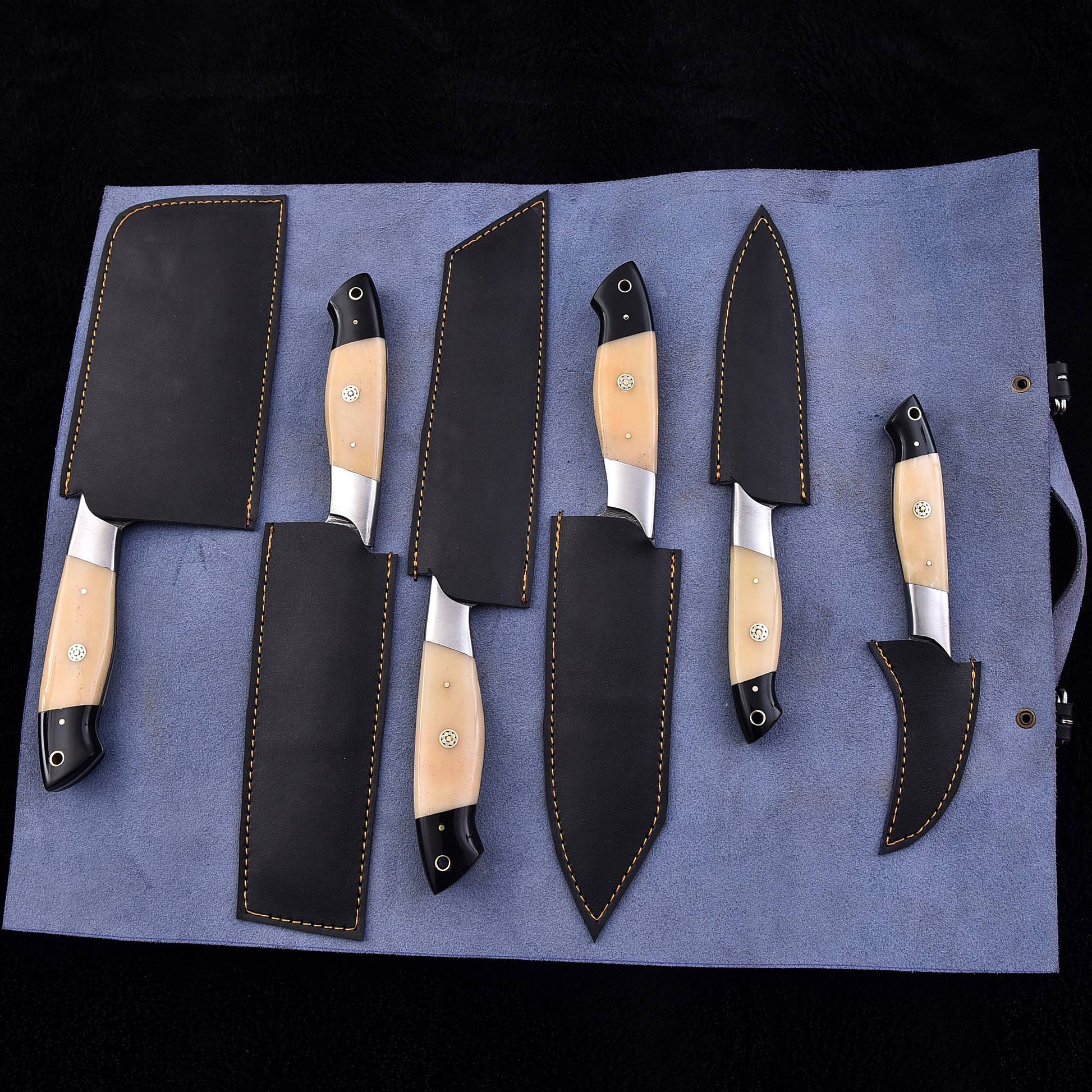 A set of six kitchen knives with light beige wood handles and black steel blades, each accompanied by a black leather sheath, arranged on a blue fabric roll against a black background.